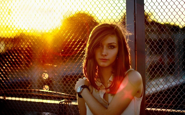 Young woman with auburn hair by a fence at sunset
