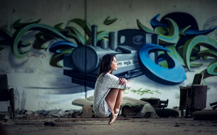 Ballerina in pointe shoes squats in front of graffiti wall