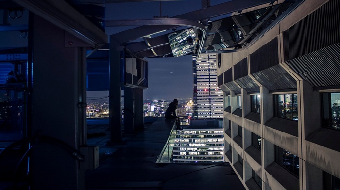 Person sitting on a skyscraper rooftop at night overlooking a cityscape