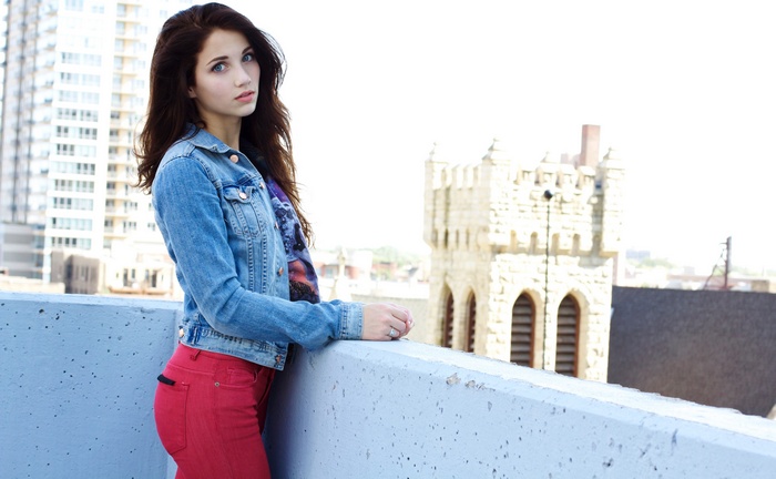 Emily Rudd on a rooftop with buildings and a tower