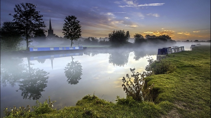 Canal boats on a misty river at sunrise with church spire