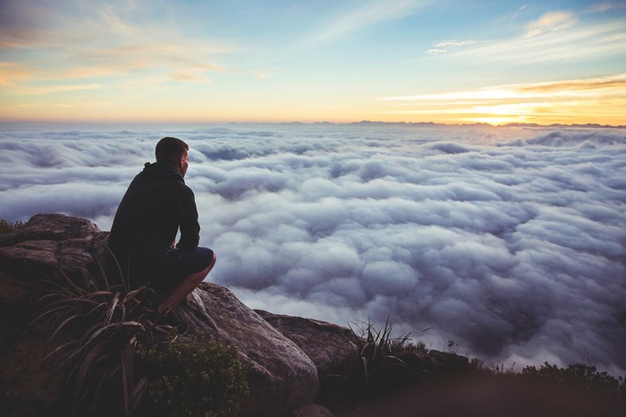 Man sitting on a cliff overlooking clouds at sunrise