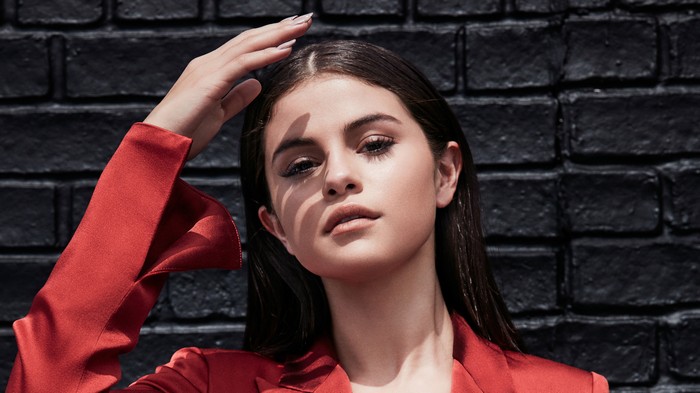 Selena Gomez in a red jacket against a brick wall
