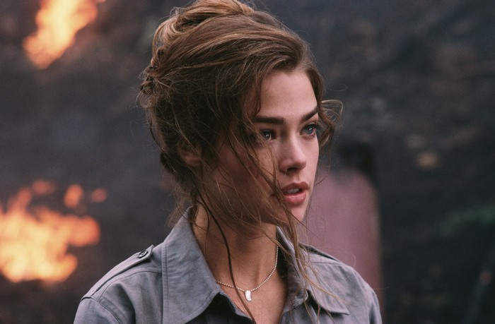 Denise Richards in a grey jacket with fire in the background