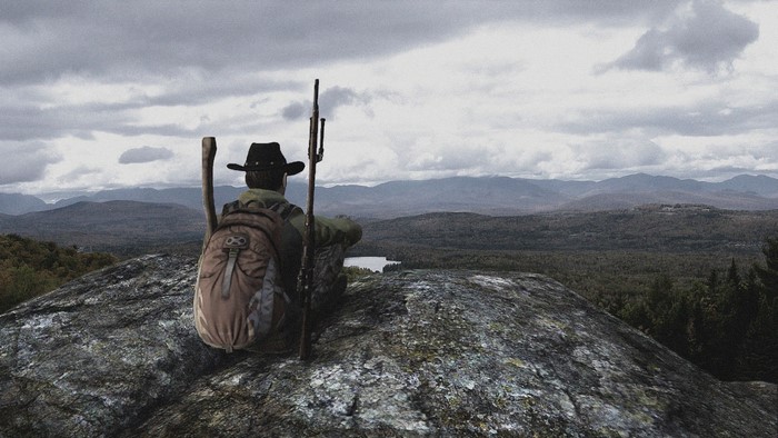 Man with backpack and rifle on mountain overlooking landscape