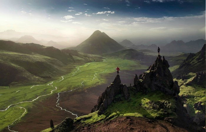 Two hikers on rocky peaks overlooking a green valley in Iceland