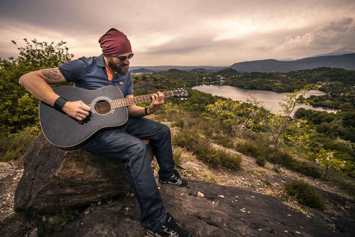 Man with beard playing guitar on a rock overlooking a lake