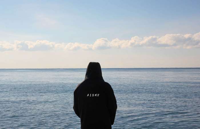 Person wearing a black hoodie with 'ALONE' text looking at the sea