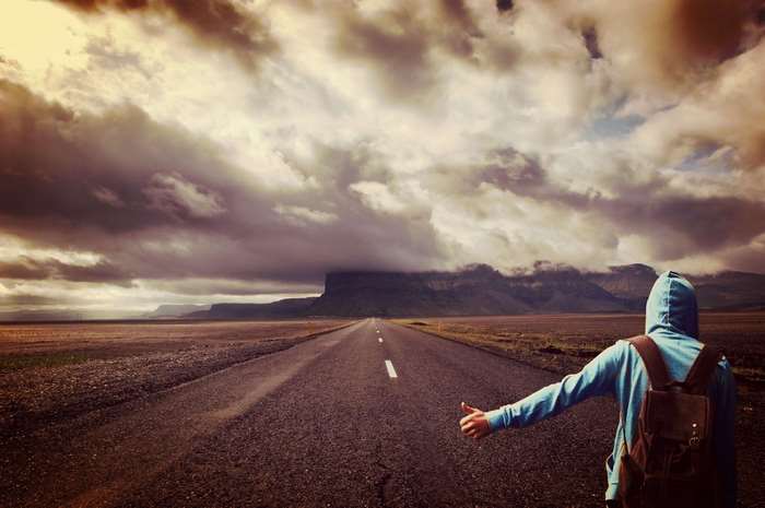 Person hitchhiking on a lonely highway under cloudy sky
