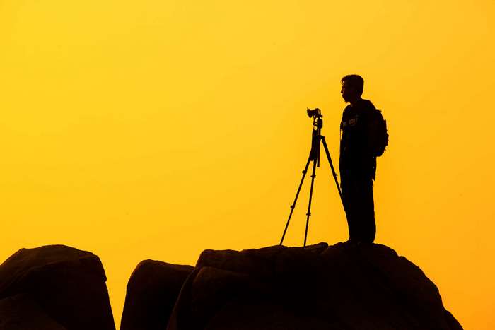 Photographer with camera on tripod on rocks, yellow sky 4K
