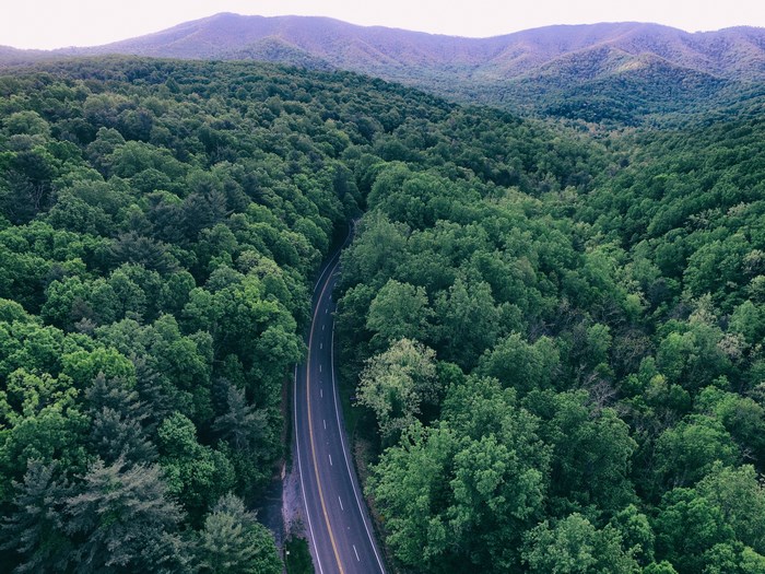Aerial view of a winding road through a dense forest with mountains in the background