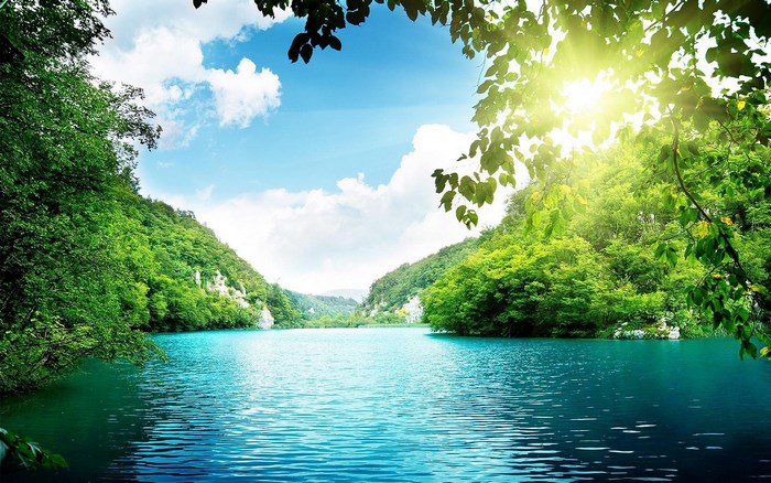 Turquoise lake surrounded by green hills and trees under a blue sky with sun rays