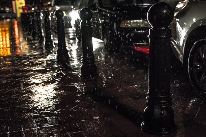 Rainy city street at night with cars and bollards