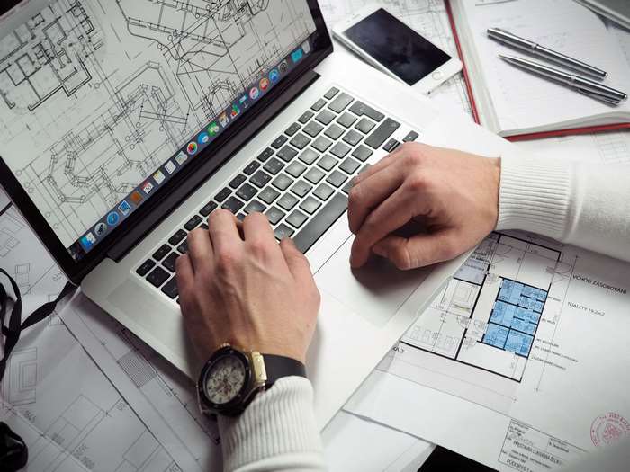 Hands typing on a MacBook with blueprints and pens