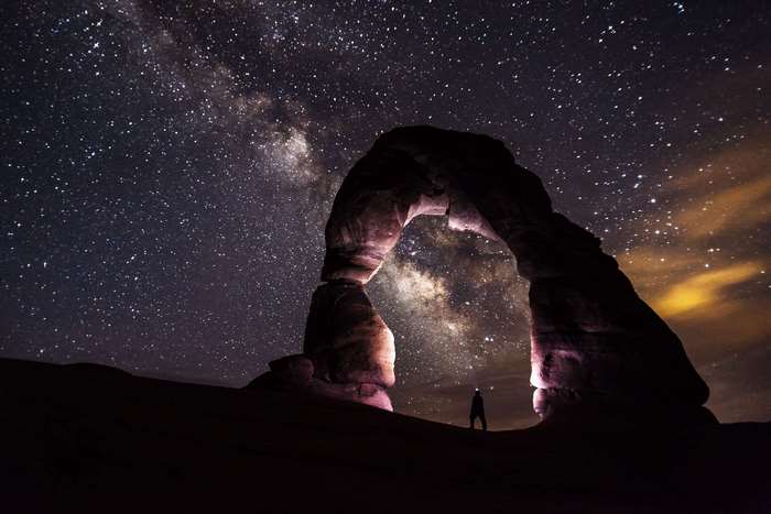 Person standing under Delicate Arch at night with Milky Way