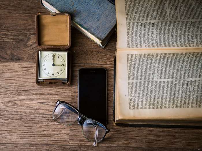 Alarm clock, smartphone, eyeglasses, and open book on wooden table
