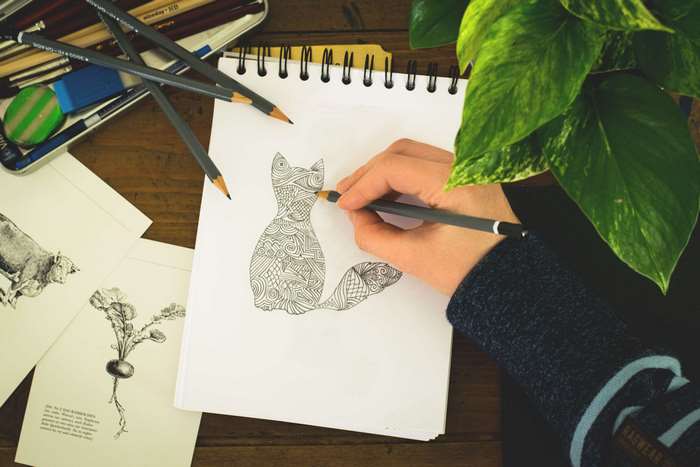 Artist drawing a patterned cat on a notebook with pencils