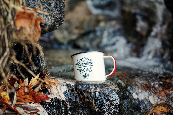 Enamel mug with "The Adventure Begins" text by a stream