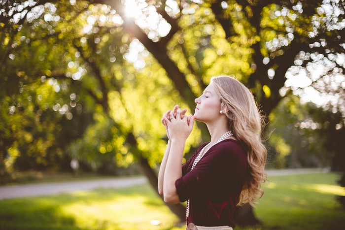 Woman praying in a park with sunlight filtering through trees
