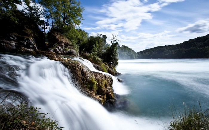 Waterfall flowing into a wide river with forested hills