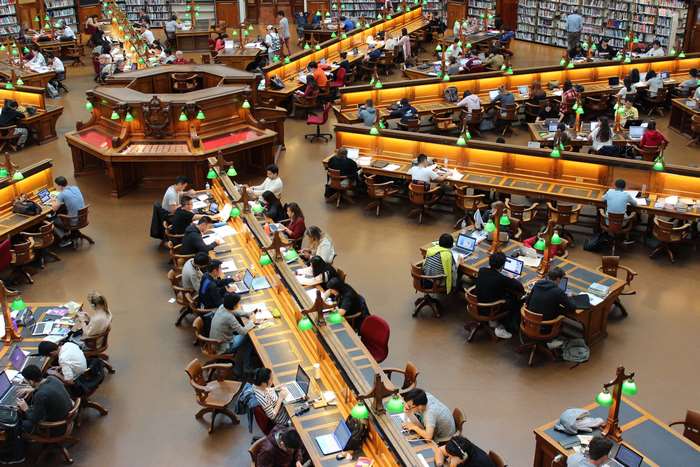 Students studying in a large library with bookshelves and tables