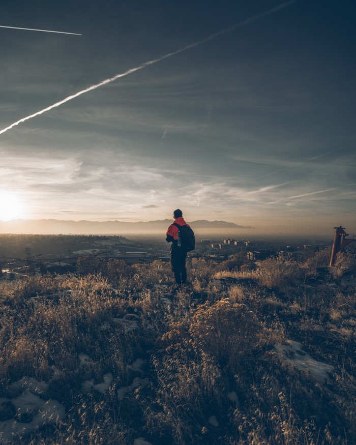 Person with backpack on grassy hill overlooking city at sunset