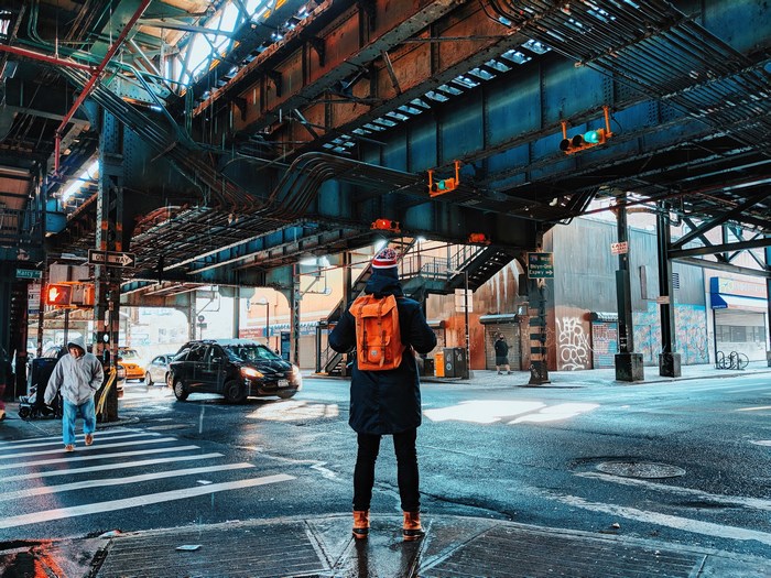 Man with orange backpack under elevated train tracks in Brooklyn
