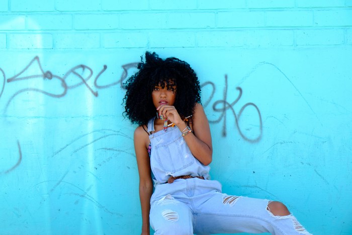 Woman in denim overalls leans against a graffiti-covered blue wall
