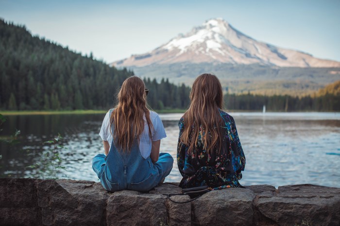 Two women sit on a stone wall overlooking a lake and mountain.
