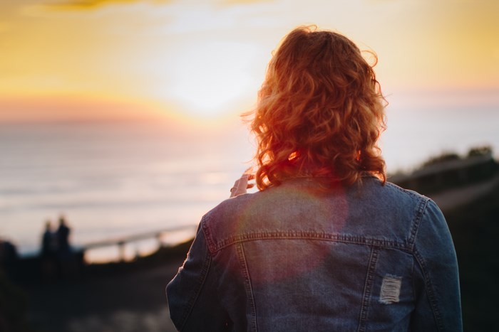 Woman with red hair in denim jacket watching sunset over ocean
