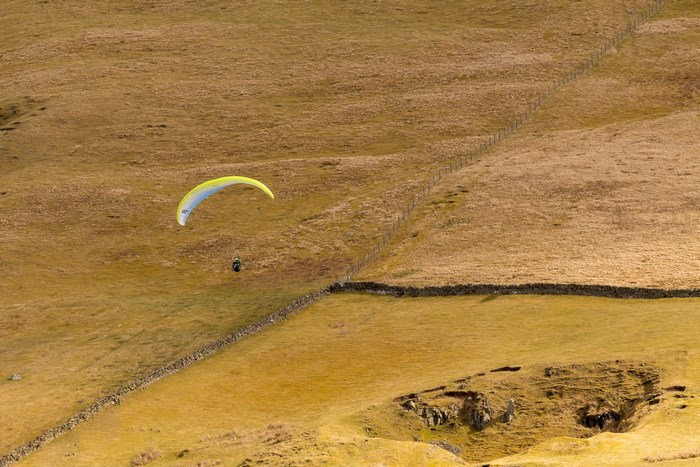 Paraglider flying over a dry, grassy field with stone walls