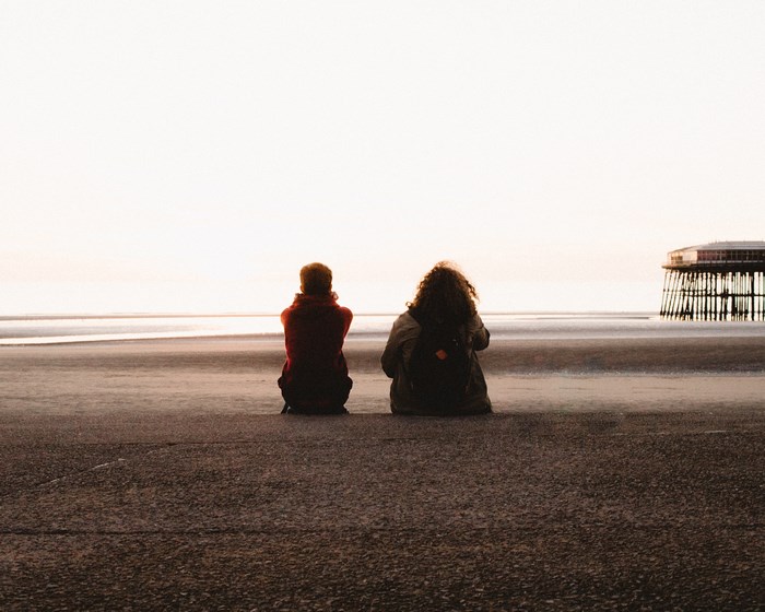 Two people sitting on a beach at sunset near a pier