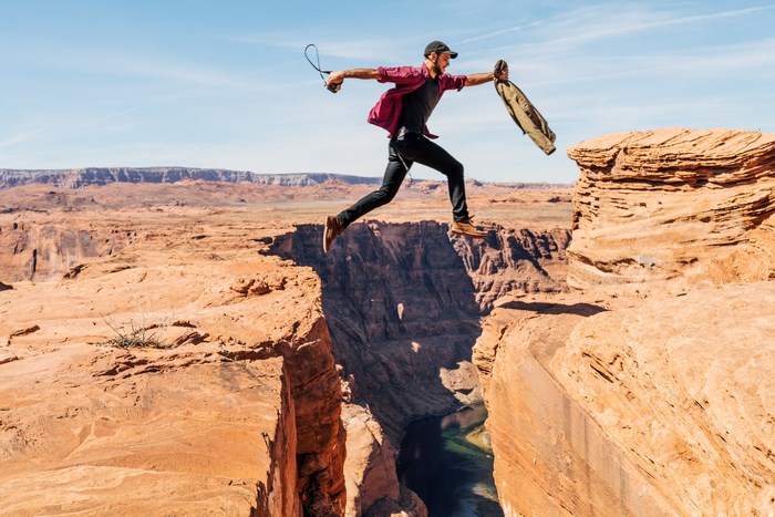 Man jumps across a canyon holding a jacket and camera