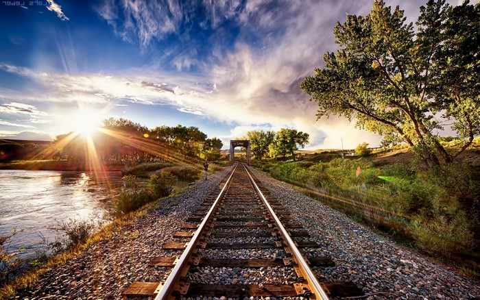 Train tracks leading to a bridge at sunset with a river