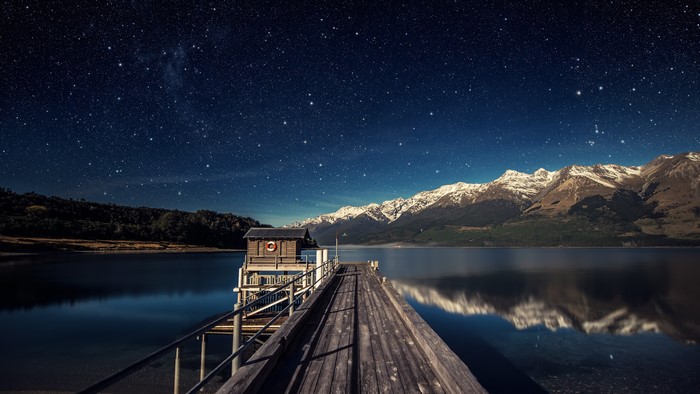 Wooden pier under starry night sky with snowy mountains
