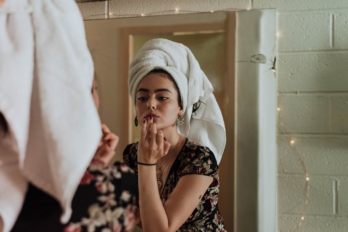 Woman applying makeup in a mirror with a towel on her head