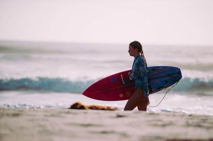 Young woman in wetsuit carrying surfboard on beach
