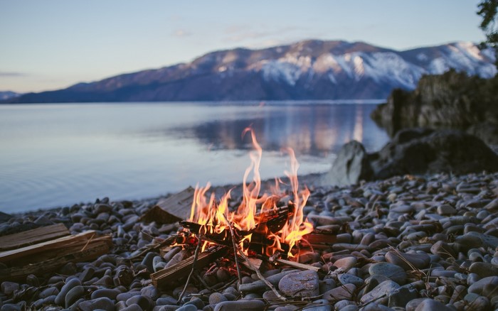 Campfire on a rocky beach by a lake with mountains