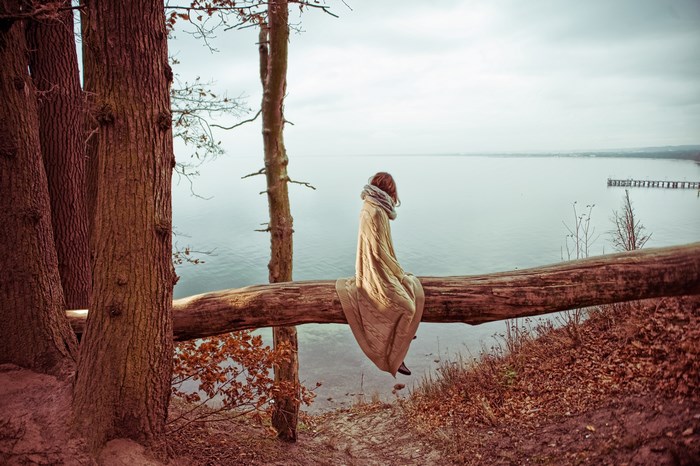 Woman wrapped in blanket sitting on a log overlooking water