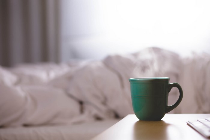 Steaming mug of coffee on a table next to an unmade bed
