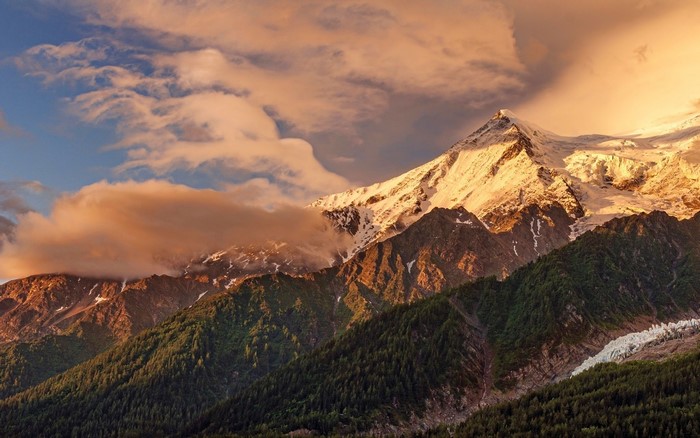 Snowy mountain peak at sunset with clouds and forest