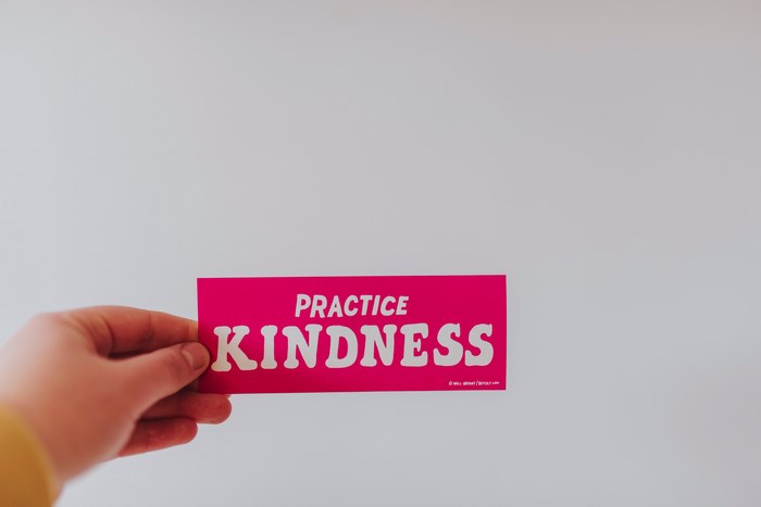 Hand holding a pink sticker with "PRACTICE KINDNESS"
