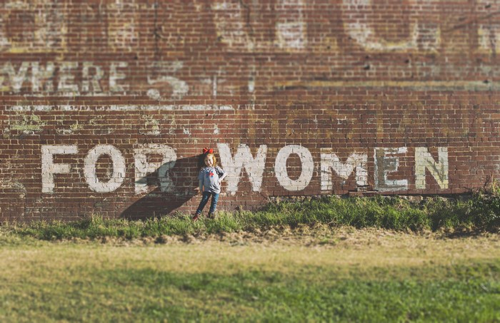 Girl in front of a brick wall with "FOR WOMEN" painted on it