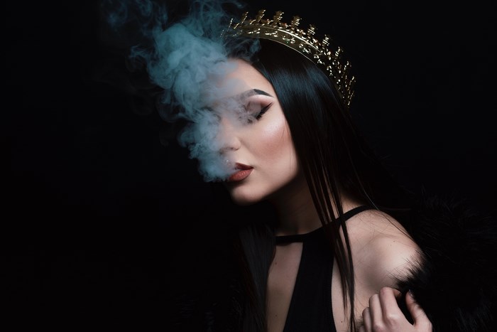 Woman with a crown exhales smoke in a dark studio