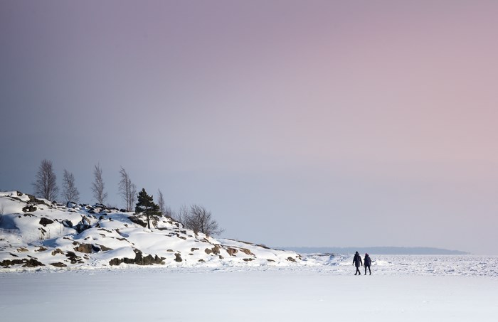 Two people walk on a frozen landscape near a rocky island in Finland