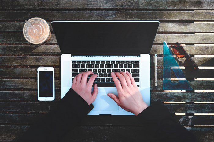 Hands typing on a MacBook with iPhone and iced coffee