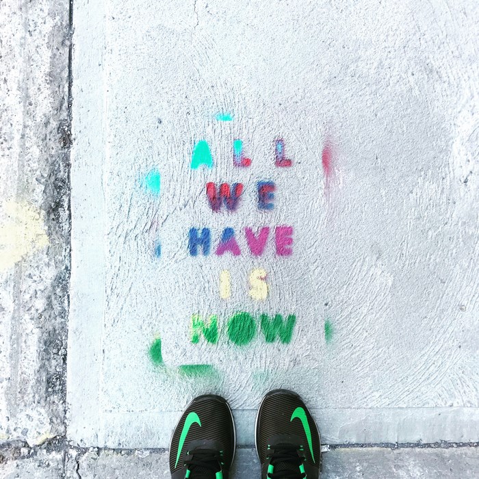 Street art quote "ALL WE HAVE IS NOW" on concrete with Nike shoes