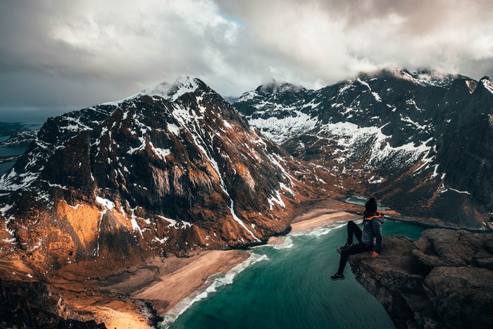 Person sitting on a cliff overlooking a snowy mountain landscape