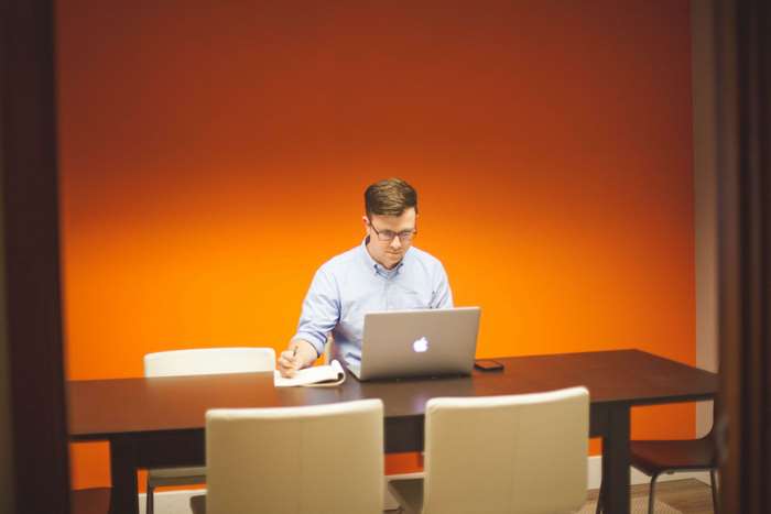 Man working on Apple MacBook at table with orange wall