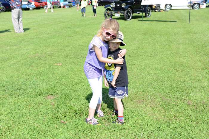 Brother and sister hugging on a grassy field with vintage cars